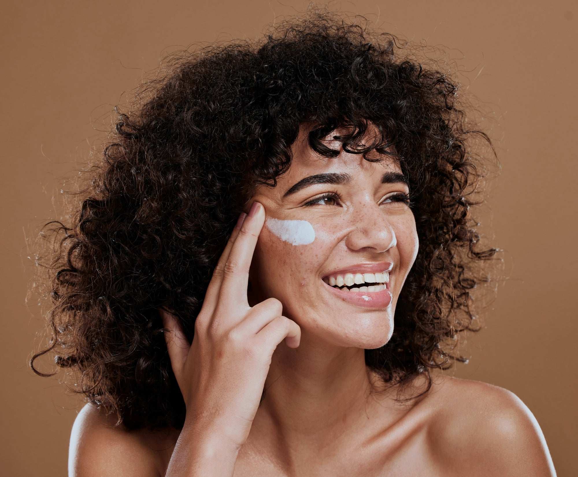 A woman with curly hair smiling and applying cream to her cheek.